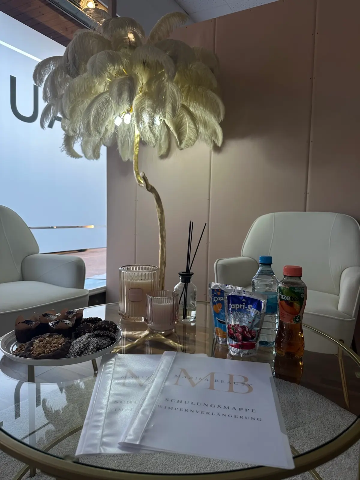 Schulungsbereich mit Sesseln, Glastisch und goldener Federlampe im Studio von Marisa Beauty